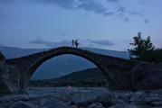 Fluss Lengariça mit Lagerfeuer, Brücke Ura e Kadiut mit Fußgängern und Berg Nemërçka in Abenddämmerung